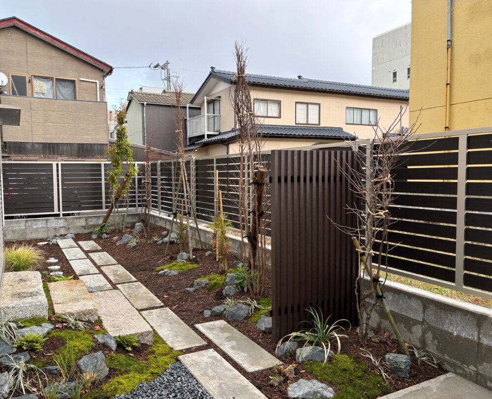 カーポートからリビング前のメインガーデンへの入口に千本格子を設け、外部とプライベート空間をやわらかく仕切りました。コンクリート土間から植栽を挟んで配置した格子越しに、奥の樹木が見え隠れすることで、庭に自然な奥行きと期待感が生まれます。