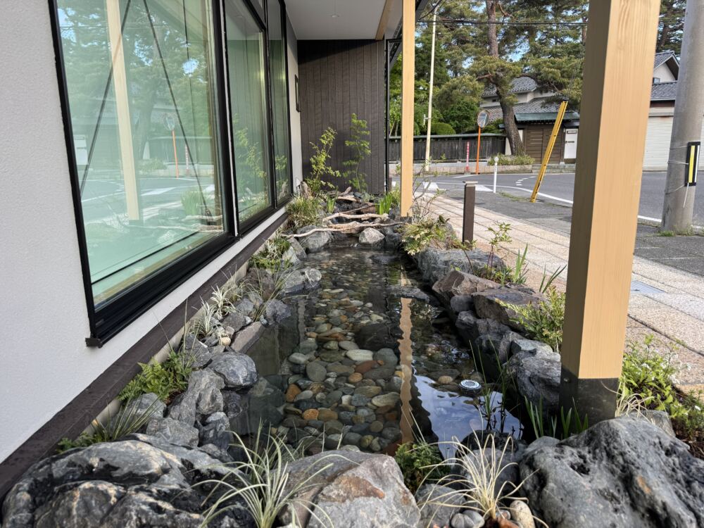 水底の美しさまでこだわった「日本の川」の再現
建物の基礎に沿って、美しいせせらぎが流れる小川をデザインしました。
大中小のゴロタ石や景石を職人の手で自然なバランスで配置し、川岸には水辺に合う植物や低木を植栽。
流木をアクセントに加えることで、まるで本物の渓谷のワンシーンを切り取ったかのような、深みのある景色に仕上げています。
ここにたくさんのメダカたちが泳ぎ回る日が今から待ち遠しくなります。
アプローチは