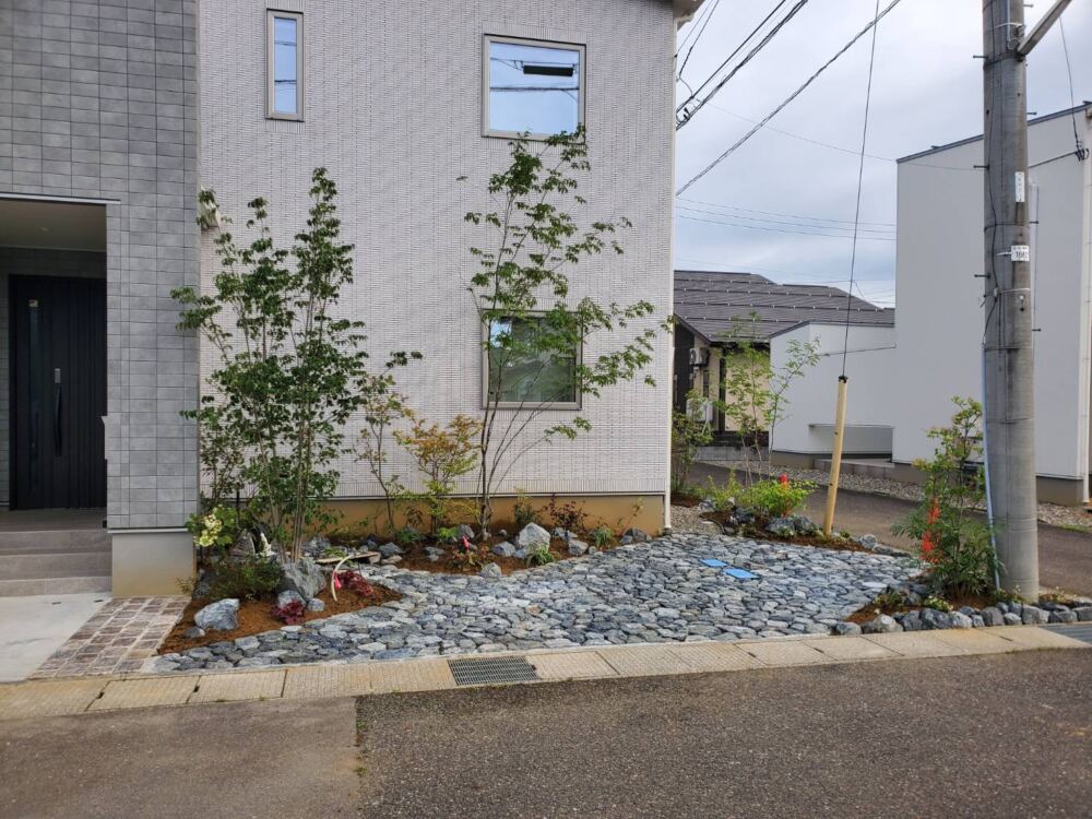 頑固な雑草を撤去して防草施工を施し、足元にはシックな表情の割栗石を美しく敷き詰めました！
普段はゴツゴツとした石の造形美が建物を引き立てつつ、「ご友人やご親戚が来られた際の臨時駐車スペース」としてフレキシブルに大活躍します。
アプローチ沿いには高木や可憐な下草を配し、雑草の手間をゼロにしながら、機能的で高級感あふれるお住まいの「顔」へと仕上げました。